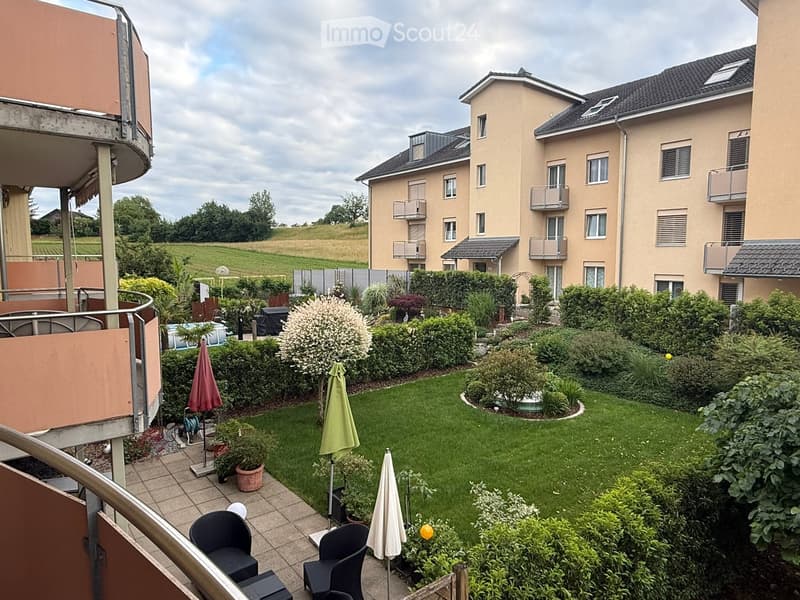 Well-maintained garden with lawn, shrubs, potted plants, patio furniture, and umbrellas. Adjacent to a multi-story residential building with balconies.