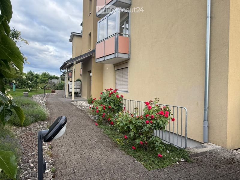 Two-story building with beige walls, a balcony with pink panels, and a pathway lined with red flowers and shrubs.