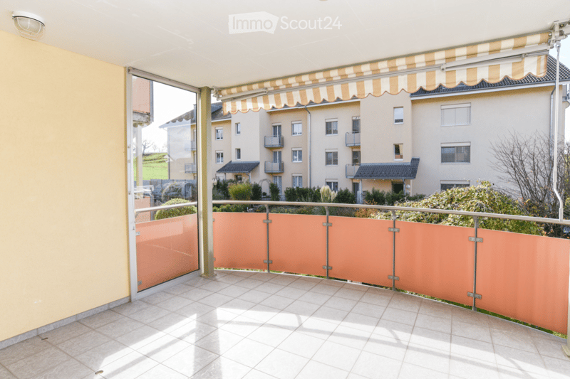 Balcony with tiled floor, metal railing, and retractable awning, overlooking a residential area with buildings and greenery.