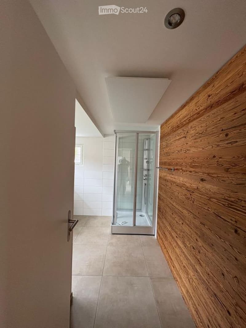 A modern hallway with a glass-enclosed shower area visible at the end, featuring light-colored tiles and a wooden wall panel on one side.