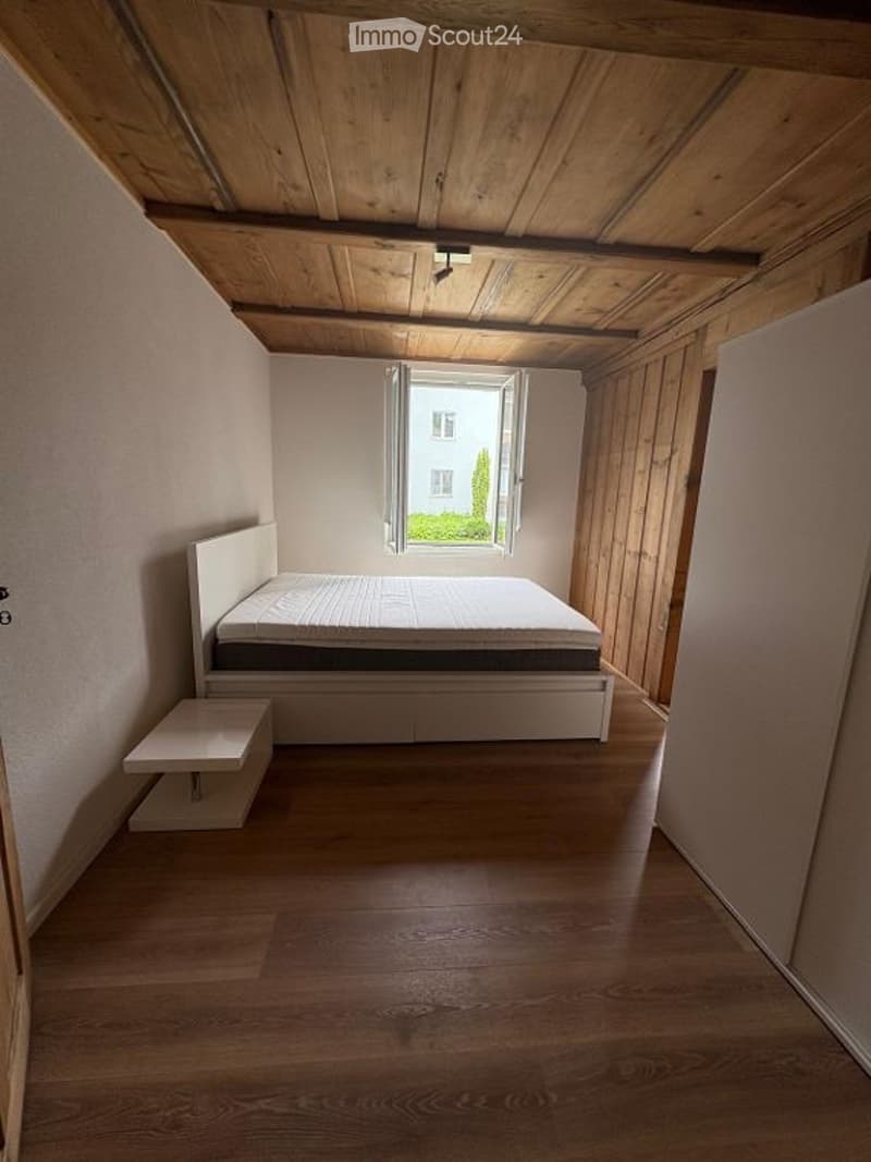 Minimalist bedroom with a white bed, nightstand, wooden floor, and exposed wooden ceiling beams. Large window with a view of greenery.