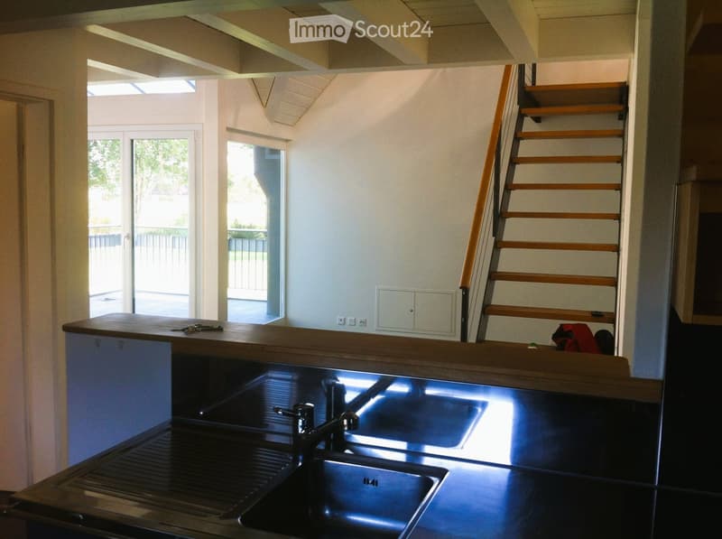 Modern kitchen with a sleek black countertop, double sink, and a wooden staircase leading to an upper level. The space features large windows and a bright, open layout.