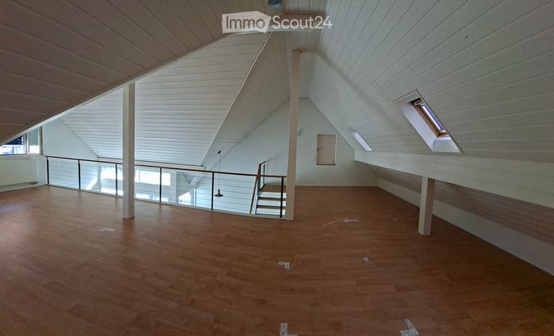 An empty room with wooden flooring, white walls, and a staircase leading to an upper level. The room features large windows and a modern, open design with exposed wooden beams on the ceiling.