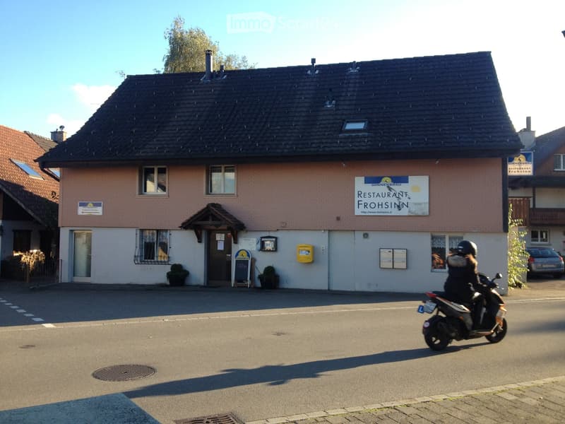 Two-story building with a restaurant sign reading "Restaurant Frohsinn" and a person riding a scooter past it.