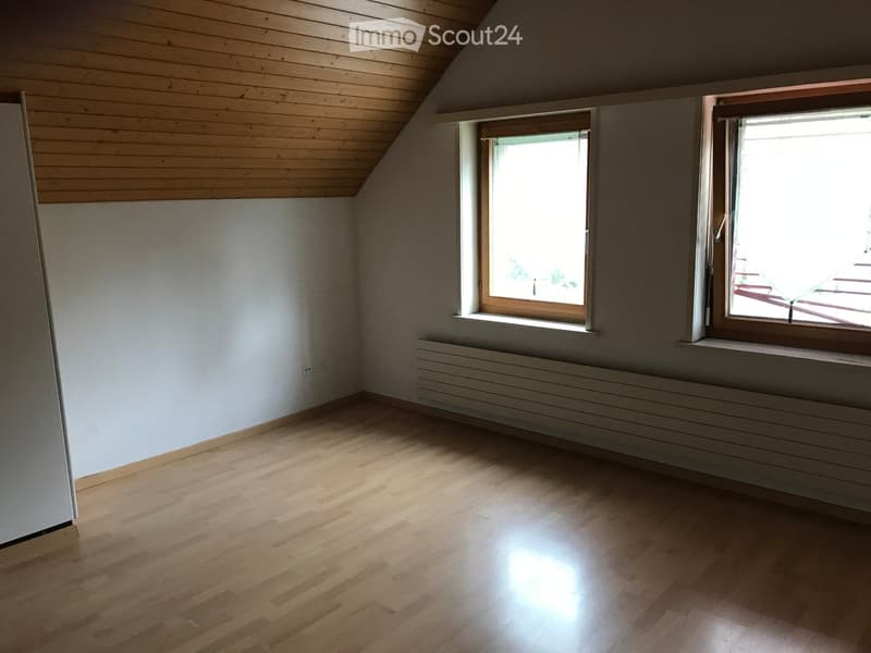 Empty attic room with wooden floors, white walls, and two windows with wooden frames. The room features a slanted wooden ceiling and a radiator below the windows.