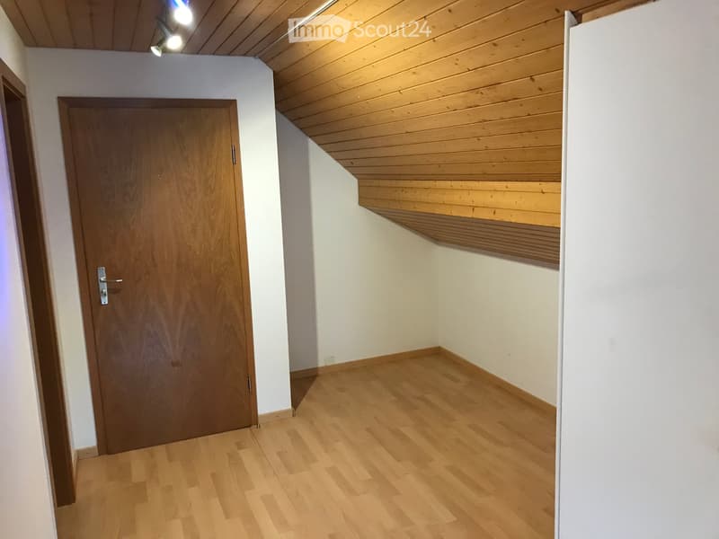 Empty room with wooden flooring, white walls, and a wooden door. The ceiling is slanted with wooden paneling and built-in shelving.