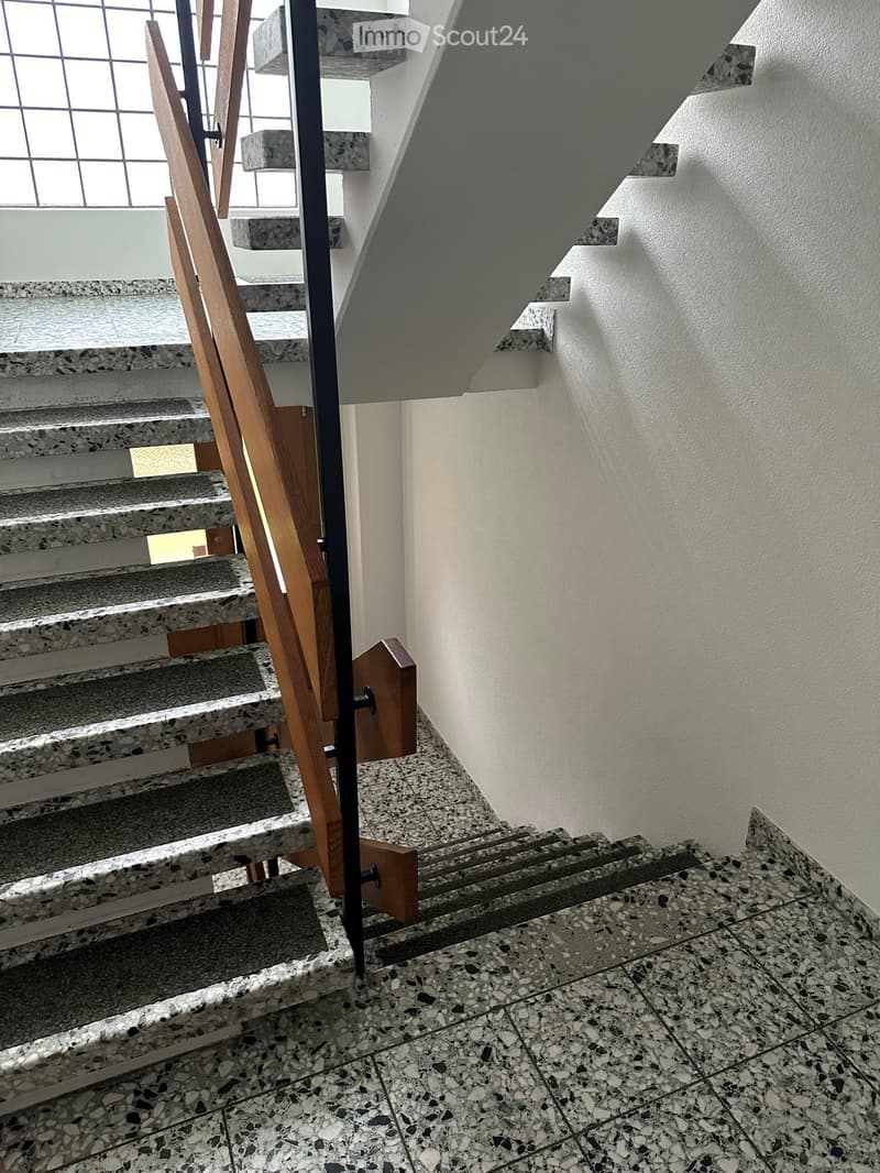 Modern staircase with wooden handrails and marble steps, leading upwards with a window on the left side.