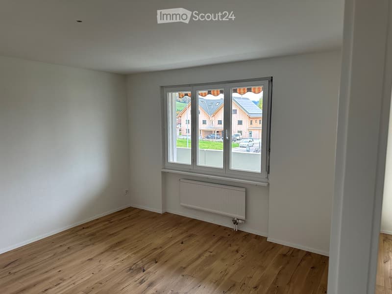 Empty room with wooden flooring, white walls, large window with view of the outside, and a radiator below the window.
