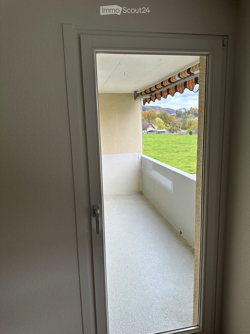 Balcony with white floor, beige walls, and a retractable awning, overlooking a green field and distant hills.
