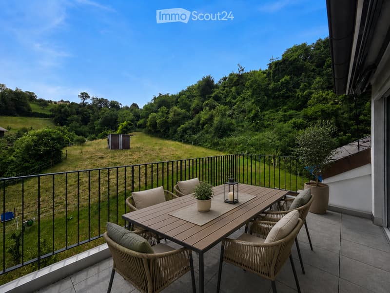 Balcony with wooden table, four wicker chairs, potted plant, and lantern, overlooking a green, hilly landscape.
