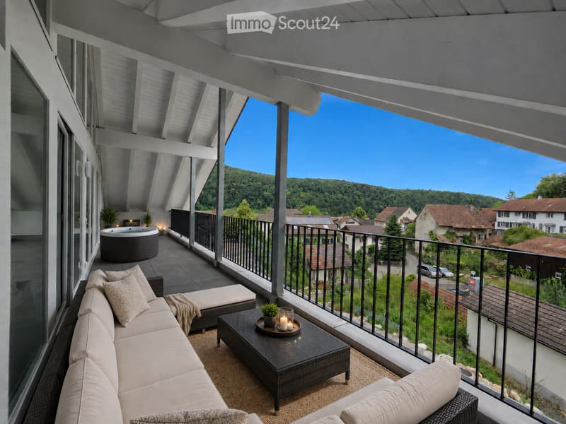 Balcony with a modern design, featuring a white wooden ceiling, a comfortable sofa, a coffee table, a hot tub, and a scenic view of a town and green hills.