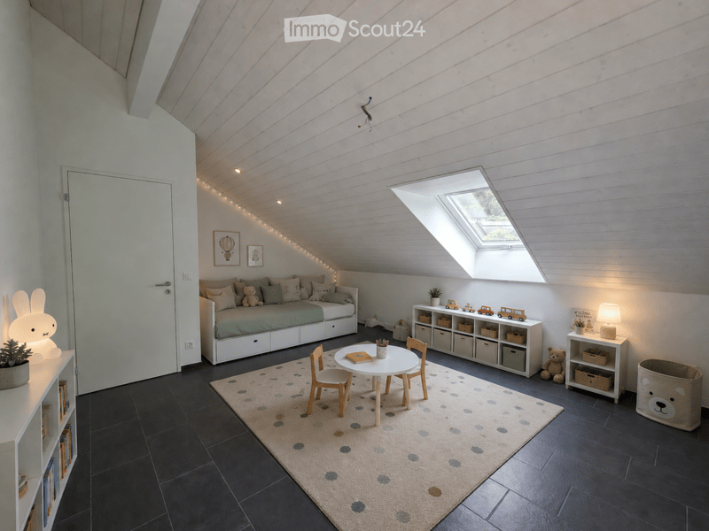 Bright, modern attic bedroom with slanted wooden ceilings, large skylight, built-in sofa bed, small round table with chairs, storage shelves, and decorative lighting.