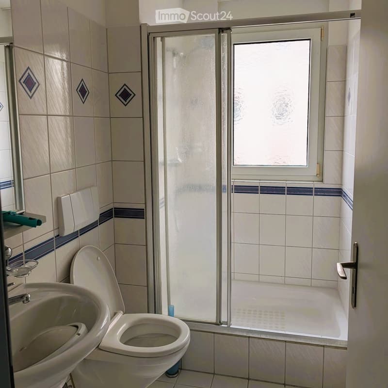 Bathroom with beige tiled walls, a toilet, a sink, a shower with a glass door, and a window above the shower. The tiles feature decorative diamond patterns and blue accents.