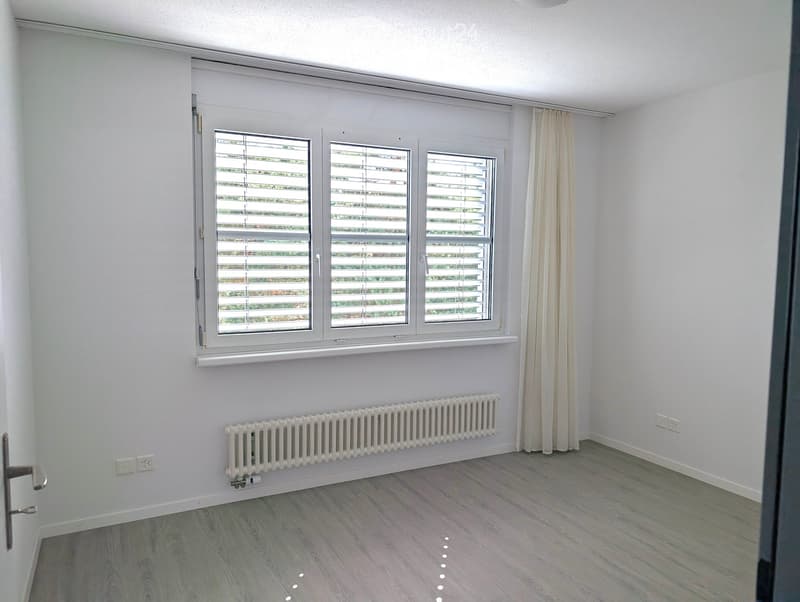 Empty room with light grey walls, wooden flooring, large windows with blinds, and a white radiator under the windows.