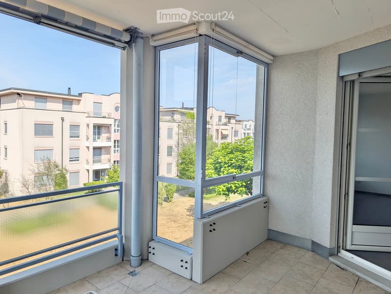 Balcony with large windows, tiled floor, metal railing, and view of surrounding buildings and greenery.
