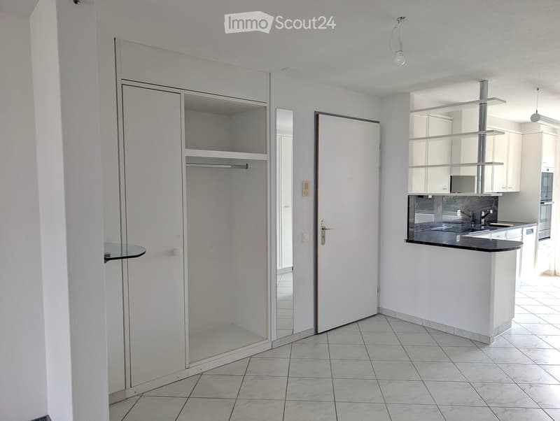 Empty room with tiled floor, white walls, and a glass table in the corner. A closet with shelves and a mirror is visible, along with a doorway leading to another room.