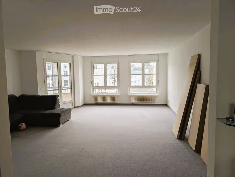 A spacious, unfurnished living room with large windows, a grey sofa, and a small ball on the floor. Wooden panels are stacked against the wall.