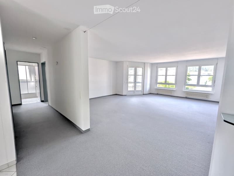 Empty room with white walls, grey carpet, large windows, and a door leading to another space.