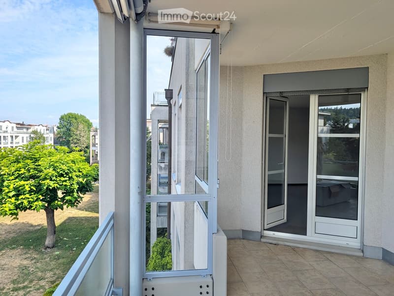 Balcony with tiled floor, glass sliding doors, and a view of a green garden area with trees and shrubs.