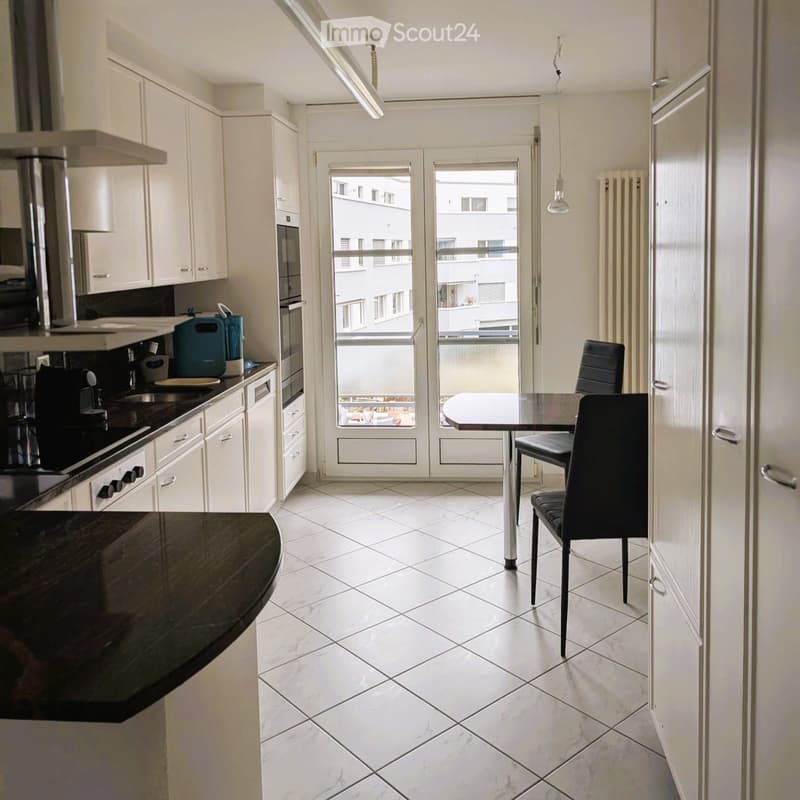 Modern kitchen with white cabinets, black countertop, and a dining area with a table and chairs. Features a large window with a view of the building outside.