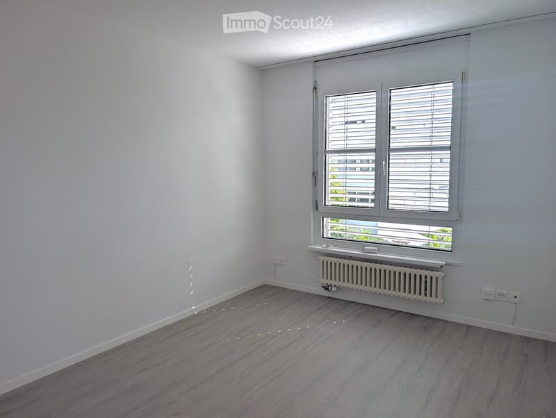 Empty room with light grey walls, wooden flooring, and a large window with blinds. A radiator is placed below the window, and electrical outlets are visible on the walls.