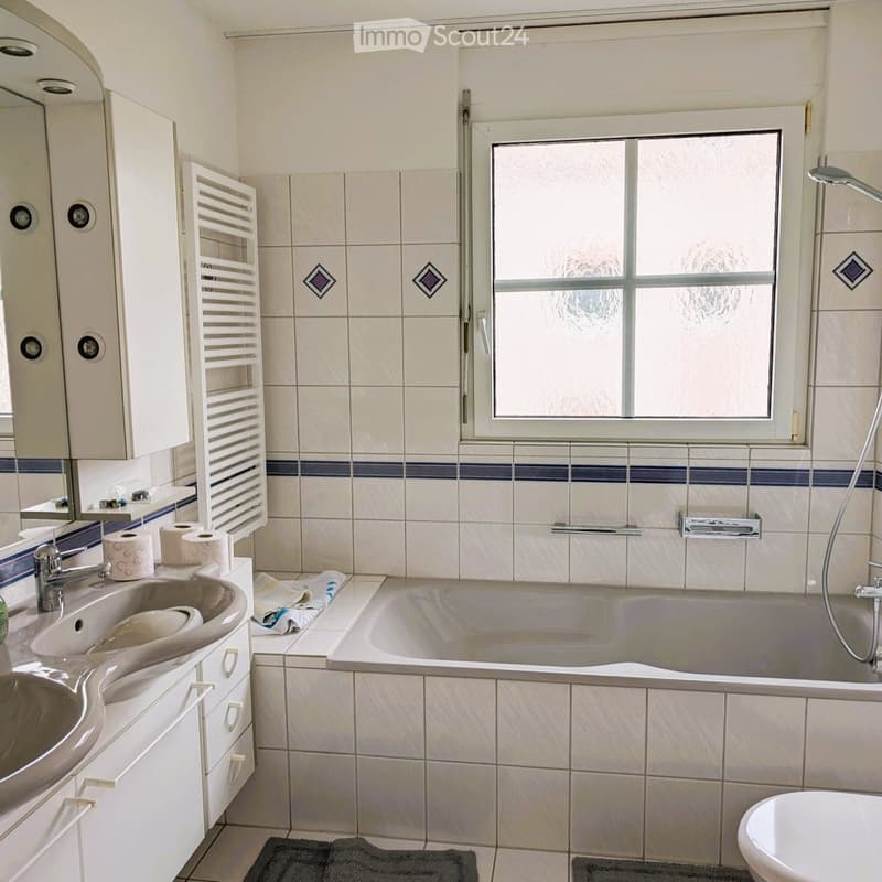 Bathroom with a sink, bathtub, toilet, mirror, and towel rack. The walls are tiled with a blue accent border, and there is a window with frosted glass.