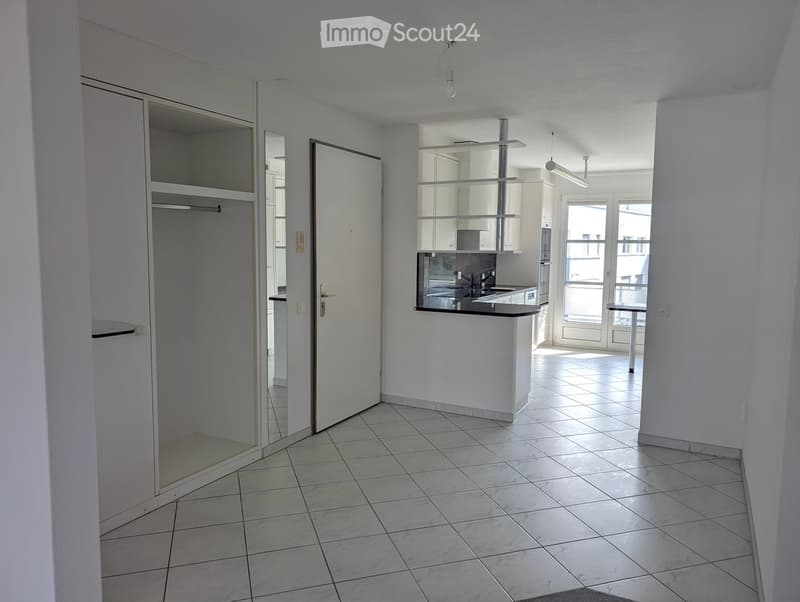 Empty room with tiled floor, white walls, and a partially visible kitchen area with cabinets and a sink.