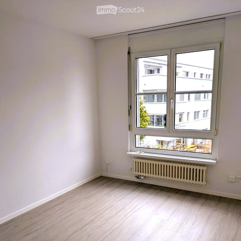 Empty room with light-colored wooden flooring, white walls, and a large window with a radiator below it. The window offers a view of neighboring buildings and a small outdoor area with tables and chairs.