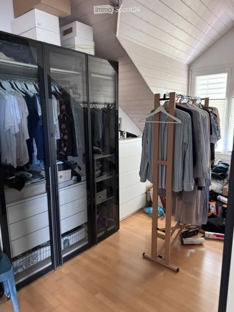A well-organized walk-in closet with a large mirrored wardrobe, a wooden clothing rack, and a small stool. The space features wooden flooring and white walls.