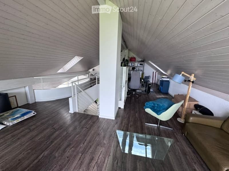 Modern, open-concept attic space with wooden flooring, slanted ceilings, and large windows. Features a sofa, glass coffee table, modern chair, floor lamp, and an office area with a desk and chair.