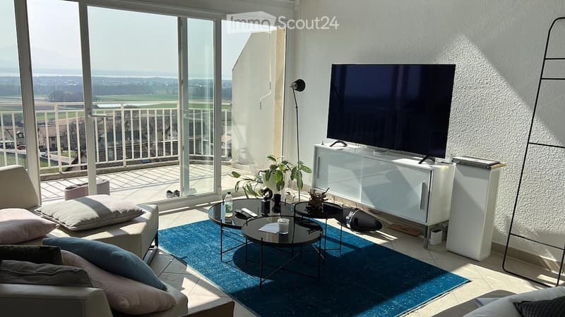 Modern living room with large windows offering a scenic view, a sofa with pillows, a coffee table, a TV stand, a floor lamp, and a small plant.