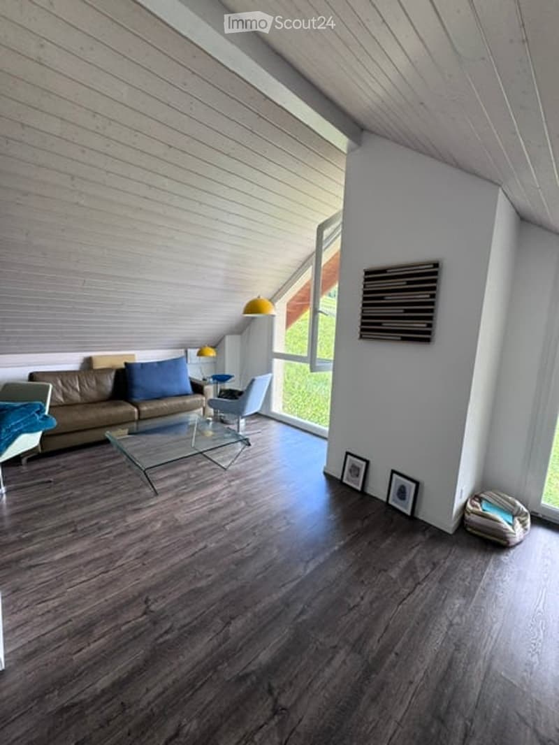 Modern living room with dark wood flooring, large windows, a brown leather sofa, a glass coffee table, a grey armchair, and a floor lamp.