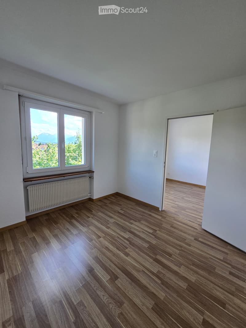 Empty room with wooden flooring, white walls, large window with view, radiator, and an open doorway leading to another space.