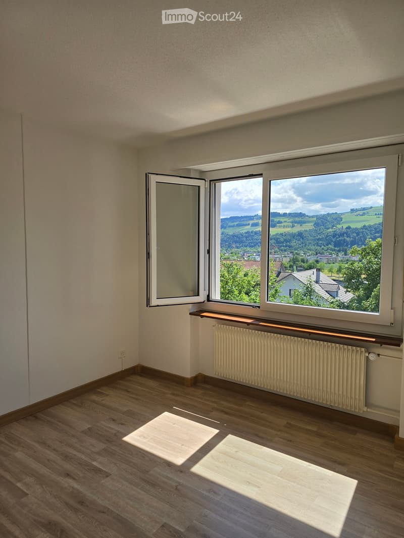 Empty room with wooden flooring, large windows offering a scenic view, radiator, and a small shelf beneath the windows.