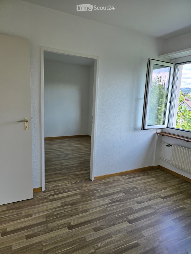 Empty room with wooden flooring, white walls, a door, and a window with a view of trees and a house outside.