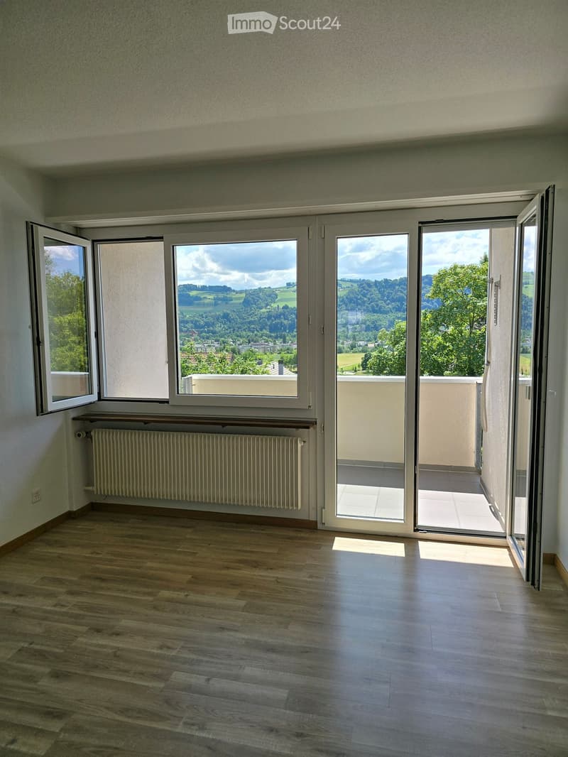 Empty room with wooden flooring, large windows opening to a balcony, radiator, and scenic view of greenery and hills.