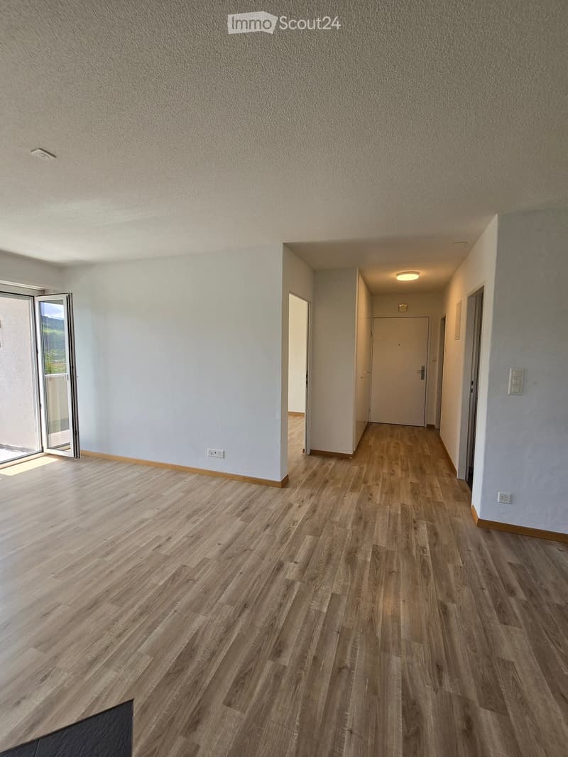 Empty hallway with wooden flooring, white walls, and recessed lighting. Features an open sliding door leading to a balcony or outdoor area on the left and a closed door at the far end.