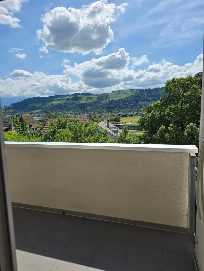 Balcony with tiled floor and beige walls, offering a scenic view of a town and surrounding hills under a partly cloudy sky.