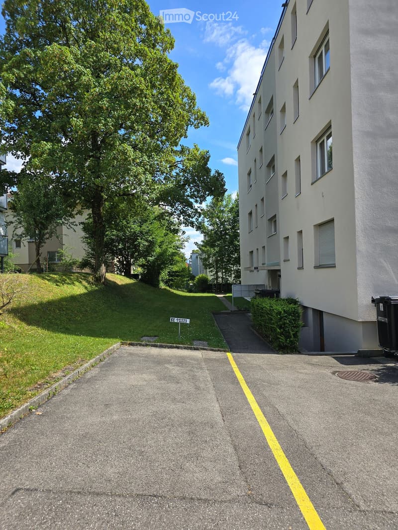 Modern multi-story building with white facade and multiple windows, situated on a raised ground floor with a paved driveway and surrounding greenery.