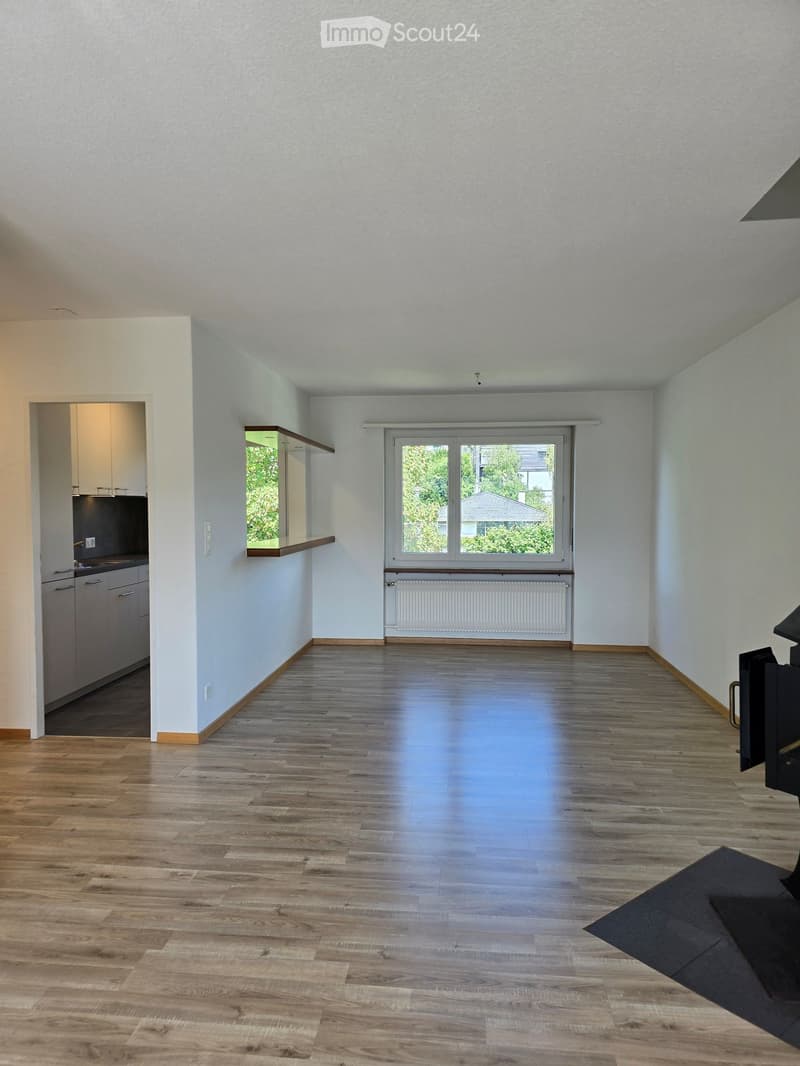 Empty room with light-colored walls, wooden flooring, and a large window with a view of greenery. A small kitchen area is visible in the background.