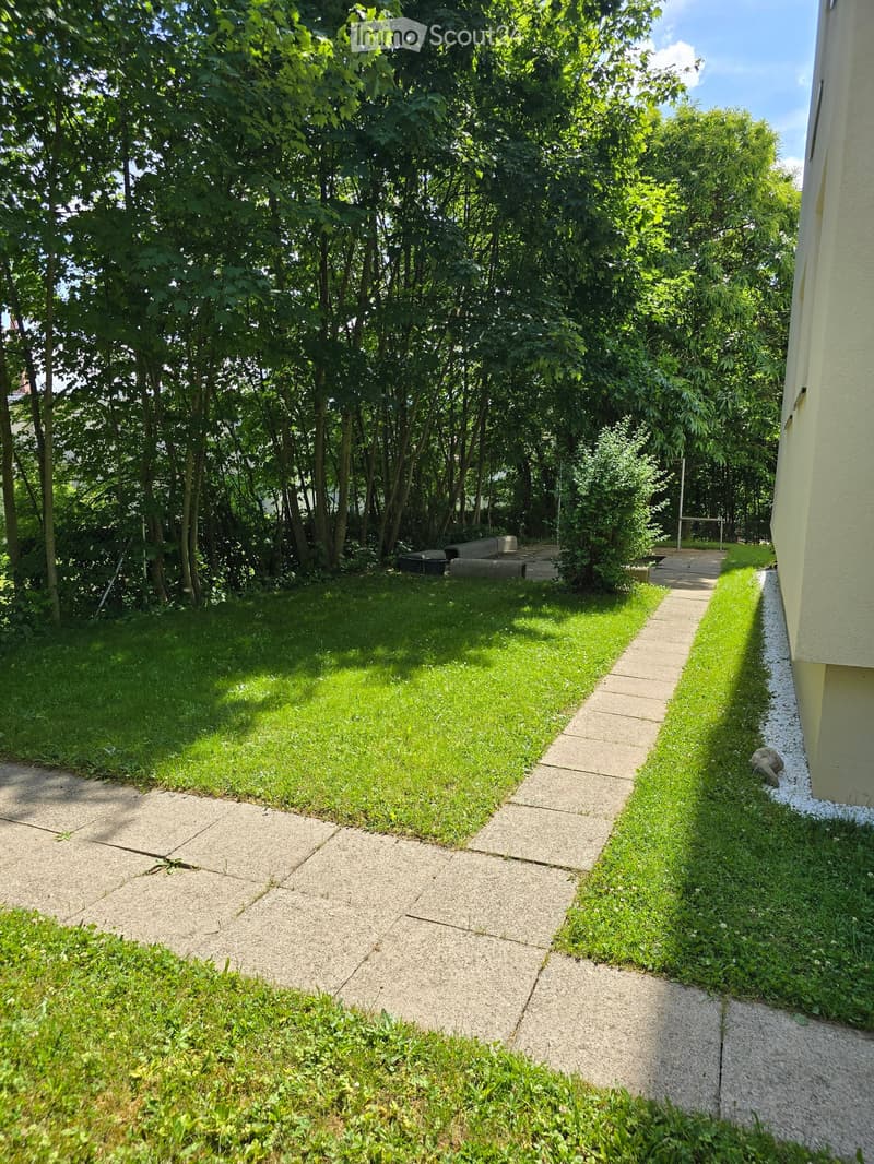A well-maintained garden with a concrete pathway, grassy areas, and surrounding trees, bordered by a building on one side.