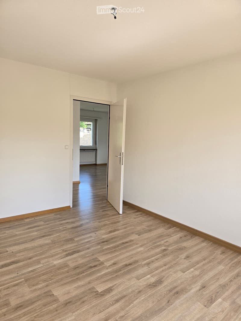 Empty room with light-colored walls and wooden flooring, featuring an open doorway leading to another room with a window and radiator visible.