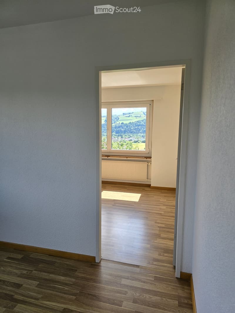 Empty hallway with wooden flooring, light-colored walls, and an open doorway leading to a room with large windows and a scenic view.