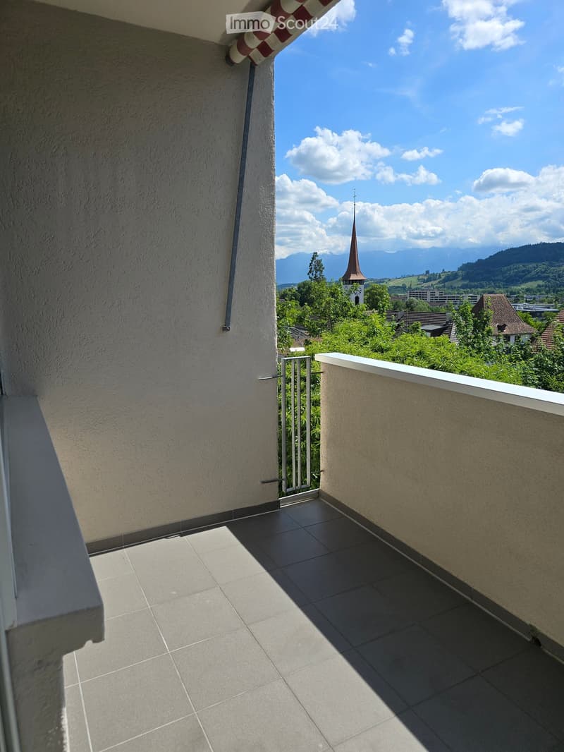 Balcony with tiled floor, metal railing, and scenic view of a town with a church spire and surrounding greenery.