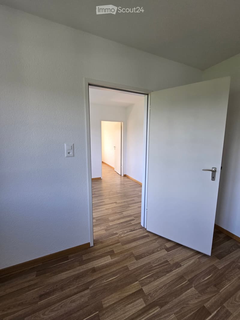Empty hallway with wooden flooring, white walls, and an open door leading to another room.