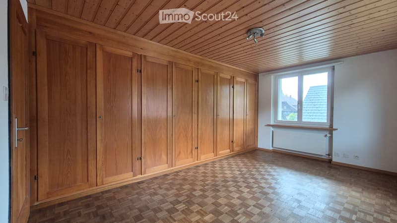 Empty room with wooden built-in wardrobes, wooden ceiling, and wooden floor. Large window with view of rooftops.