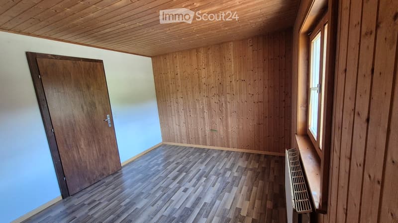 Empty room with wooden walls, wooden floor, and a single window with a radiator below it. The room has a wooden door on the left side.