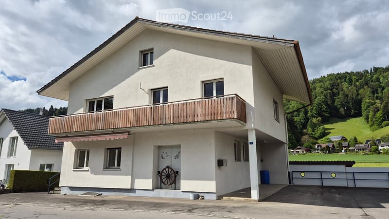 Two-story house with white exterior, large windows, and a wooden balcony on the upper level. The house features a garage and is situated in a rural area with green hills in the background.