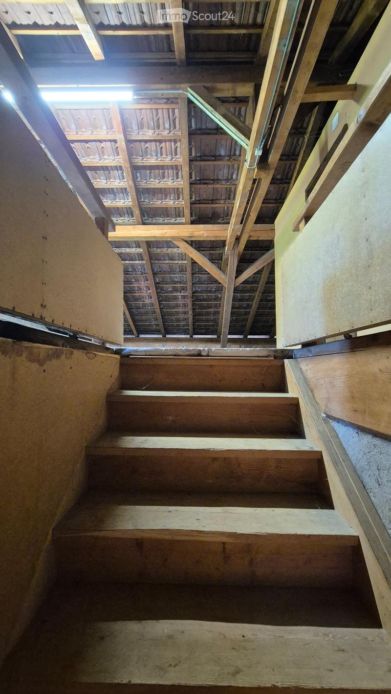 Wooden staircase with exposed wooden beams and a metal railing, leading to an open attic space with visible wooden ceiling beams and natural light coming through a skylight.