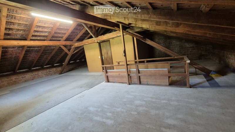 Empty attic space with exposed wooden beams, concrete floor, and a wooden staircase leading to another area.